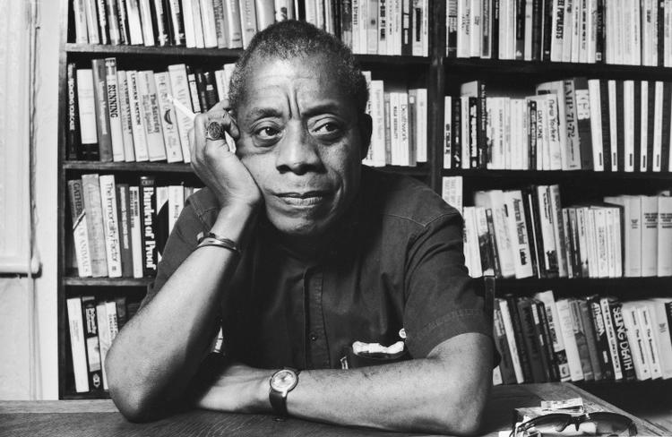 Author James Baldwin, an African American man, at a desk in front of a shelf full of books.